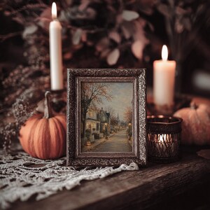 May include: A rectangular picture frame with an ornate metallic finish containing a painting of a quiet street scene with trees, houses, and street lamps. The frame is surrounded by two lit white candles, two orange pumpkins, and a metal candle holder.