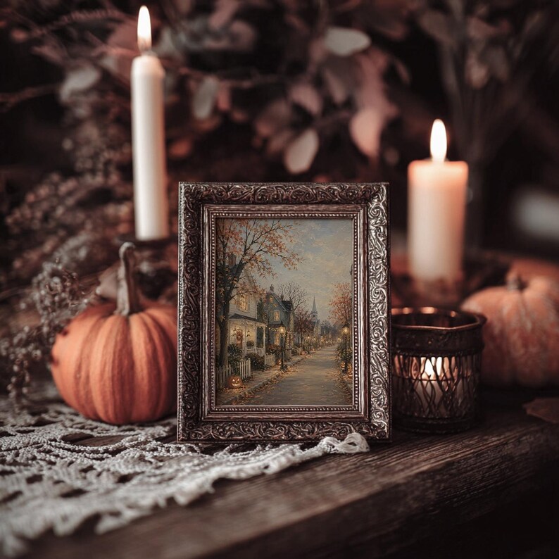 May include: A rectangular picture frame with an ornate metallic finish containing a painting of a quiet street scene with trees, houses, and street lamps. The frame is surrounded by two lit white candles, two orange pumpkins, and a metal candle holder.
