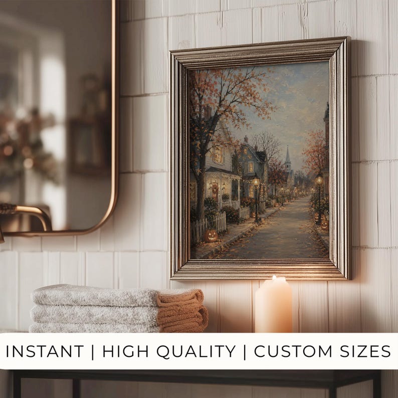 May include: A framed wall art print depicting a quiet autumn street scene at dusk with houses, glowing street lamps, and pumpkins. The artwork is displayed in a silver frame on a white tiled wall above a stack of folded towels and a pillar candle.