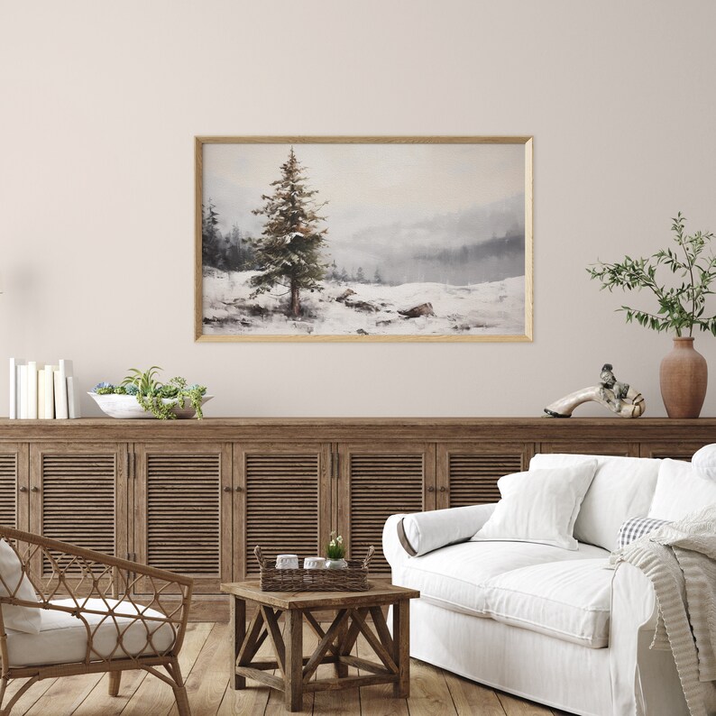 May include: A watercolor painting of a snowy landscape with a single evergreen tree in the foreground. The painting is framed in a light wood frame and hangs on a white wall above a wooden cabinet.