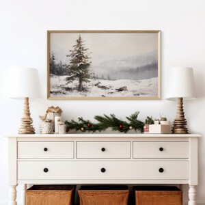 May include: A framed watercolor painting of a snowy winter scene with a single pine tree in the foreground. The painting is hanging on a white wall above a white dresser with three drawers. A green garland with red and gold ornaments is draped across the top of the dresser.