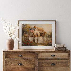 May include: A framed painting of a white cottage with a red, white, and blue American flag waving in the wind. The cottage is surrounded by a white picket fence and a garden of red and white flowers.
