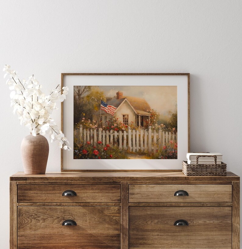 May include: A framed painting of a white cottage with a red, white, and blue American flag waving in the wind. The cottage is surrounded by a white picket fence and a garden of red and white flowers.