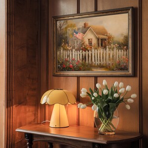 May include: A yellow table lamp with a mushroom-shaped shade sits on a wooden table next to a vase of white tulips. A framed painting of a house with an American flag hangs on the wall behind the lamp.