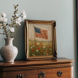 May include: A framed painting of the American flag waving in a field of wildflowers. The painting has a gold ornate frame and is displayed on a wooden dresser. A white vase with white flowers is to the left of the painting.