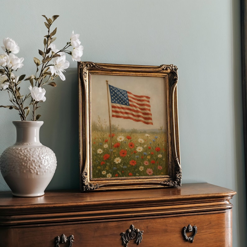 May include: A framed painting of the American flag waving in a field of wildflowers. The painting has a gold ornate frame and is displayed on a wooden dresser. A white vase with white flowers is to the left of the painting.