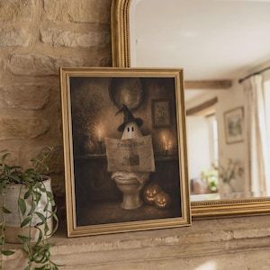 May include: A framed wall art print featuring a ghost wearing a black witch hat, sitting on a toilet while reading a newspaper titled Evening Howl. Two lit jack-o'-lanterns sit on the floor beside the ghost in a dimly lit, moody interior setting.