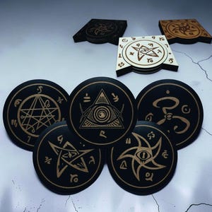 May include: A collection of circular and square coasters featuring engraved occult symbols. The circular coasters are black with gold-toned etched designs, including pentagrams and an all-seeing eye. The square coasters are black, white, and wood-toned.