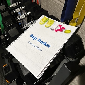 May include: A spiral-bound "Rep Tracker" notebook with the words "Rep Tracker" and "Corrective Edition" in blue. The notebook is decorated with yellow sneakers, a pink airplane, and a green oval. The notebook is resting on a set of dumbbells.