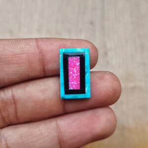 May include: A rectangular turquoise gemstone with a black border and a pink, glittery center. The gemstone is held in a hand, showcasing its vibrant colors and intricate design. This unique piece could be used for jewelry making.