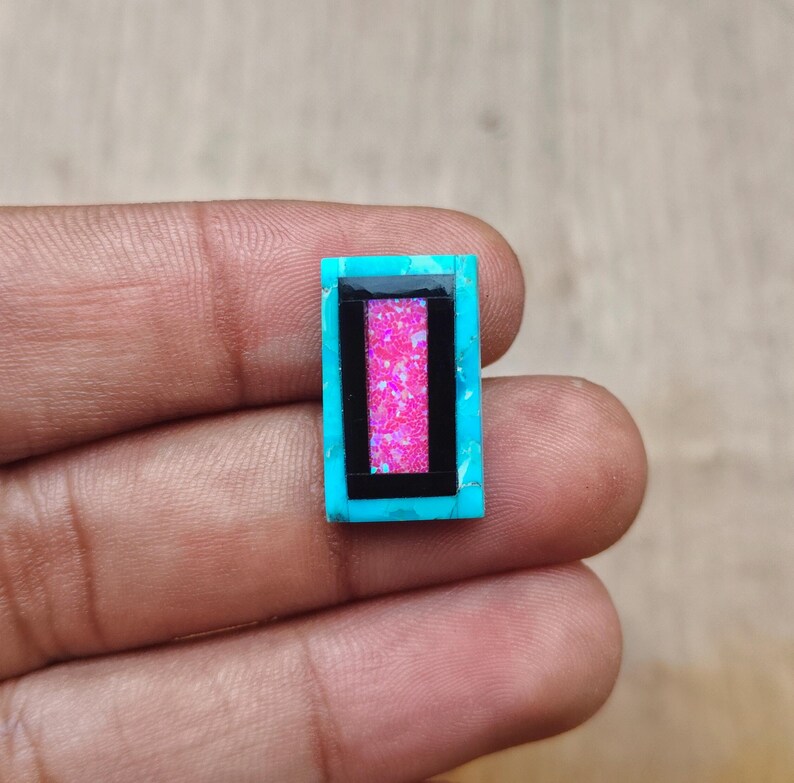 May include: A rectangular turquoise gemstone with a black border and a pink, glittery center. The gemstone is held in a hand, showcasing its vibrant colors and intricate design. This unique piece could be used for jewelry making.
