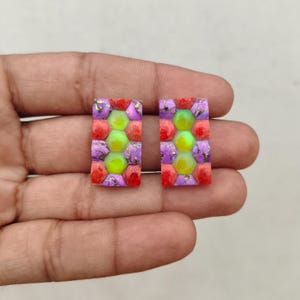 May include: A pair of rectangular earrings featuring a mosaic pattern of hexagonal lime green stones surrounded by purple and red stones. The stones are arranged in a grid-like formation, creating a vibrant, multi-colored geometric design.