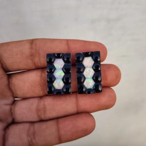 May include: A pair of rectangular stud earrings featuring a honeycomb-inspired pattern. Each earring contains three central iridescent white opal-like stones surrounded by a border of smooth, glossy black circular accents in a grid arrangement.