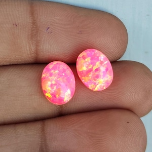 May include: Two oval-shaped gemstones with a vibrant pink hue and internal flashes of yellow and orange. The gemstones are held in a hand, showcasing their polished surfaces and potential for jewelry making or collecting.
