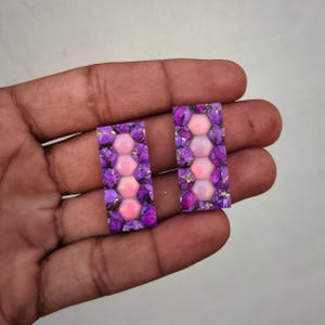 May include: Two rectangular cabochons featuring a central vertical row of four smooth, light pink hexagonal stones surrounded by a vibrant purple, marbled matrix with thin, metallic gold-colored veins embedded throughout the stone surface.