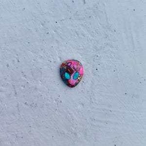 Può includere: Un piccolo cabochon rotondo in resina con un viso alieno rosa e occhi turchesi.
