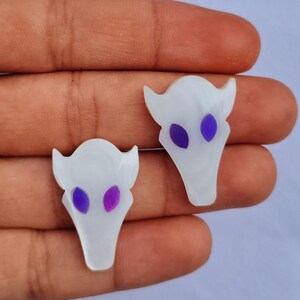 以下が含まれることがあります： 紫と青の目をした白いレジン製の頭蓋骨チャーム2個。頭蓋骨は角と尖った底を持っています。