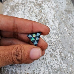 Könnte beinhalten: Ein dreieckiger schwarzer Opal-Cabochon mit einem geometrischen Muster aus kleineren Dreiecken in schillernden Farben von Grün, Blau und Gold.