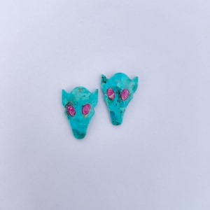 May include: Two turquoise-colored, abstract-shaped earrings with pink eye details. The earrings have a textured appearance and are set against a white background. The design resembles a stylized animal or creature.