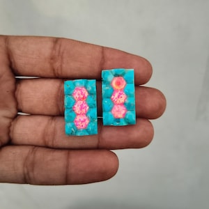 May include: Two rectangular jewelry inlays featuring a central vertical row of three hexagonal pink iridescent stones surrounded by a border of bright turquoise blue stones. The pieces have a smooth, polished finish and a geometric design.