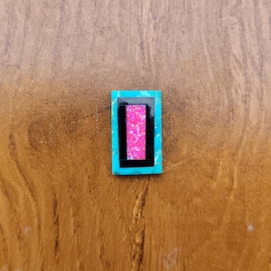 May include: A rectangular turquoise-colored gemstone with a black and pink inlay. The pink inlay is a rectangle within a black rectangle. The gemstone is set against a brown wooden surface.