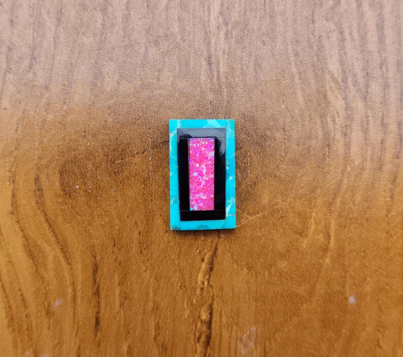 May include: A rectangular turquoise-colored gemstone with a black and pink inlay. The pink inlay is a rectangle within a black rectangle. The gemstone is set against a brown wooden surface.