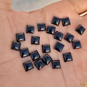 May include: A scattering of small, square, black gemstones with a polished surface. The gems are displayed on a hand, highlighting their shape and colour. The gemstones appear to be approximately 0.6 cm in size.