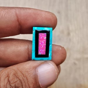 May include: A rectangular gemstone with a turquoise border, a black inner frame, and a vibrant pink center. The gemstone is held between fingers, showcasing its polished surface and color contrast. The design is geometric and eye-catching.