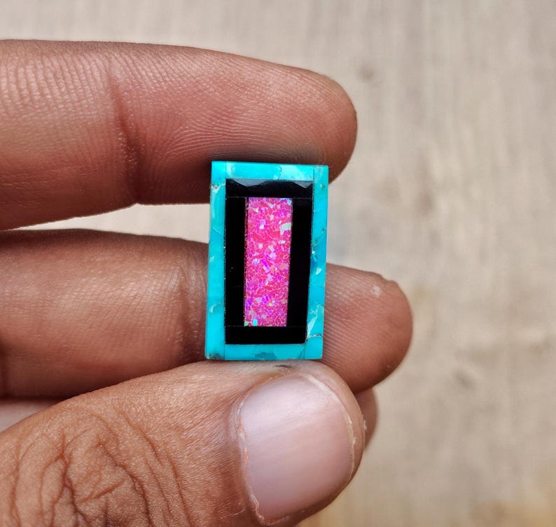 May include: A rectangular gemstone with a turquoise border, a black inner frame, and a vibrant pink center. The gemstone is held between fingers, showcasing its polished surface and color contrast. The design is geometric and eye-catching.