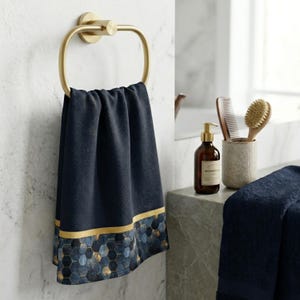 Pode incluir: Uma toalha azul marinho com uma borda dourada e com padrão geométrico pendurada em um porta-toalhas dourado. A toalha é exibida em um ambiente de banheiro, sugerindo um foco na decoração e acessórios de banheiro.