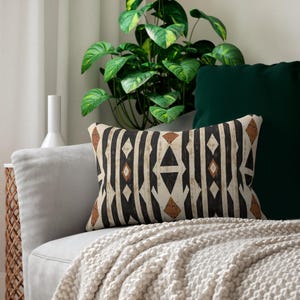 May include: Rectangular decorative pillow with a geometric pattern in black, brown, and cream. The pillow features vertical stripes and diamond shapes. It is placed on a gray sofa with a cream-colored blanket and a green pillow.