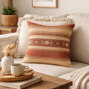 May include: A decorative throw pillow with a striped pattern in shades of beige and rust. The pillow features a central band with geometric designs. It rests on a neutral-toned sofa, suggesting a cozy living room setting.