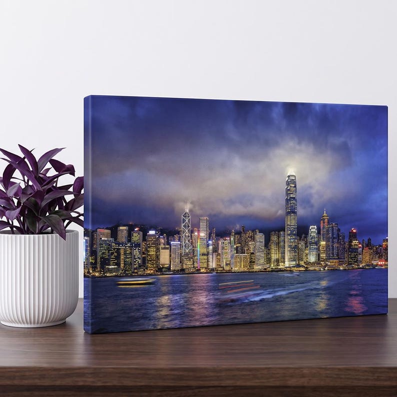May include: A rectangular canvas print displaying a night cityscape of Hong Kong across a harbor, featuring illuminated skyscrapers under a dark, cloudy sky with light trails from boats on the water, positioned next to a potted purple plant.