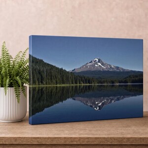 May include: A rectangular canvas print featuring a landscape of a snow-capped mountain peak rising above a dense forest of dark green pine trees, with the entire scene reflected clearly in the calm, dark blue water of a lake below.