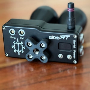 Op de afbeelding: Een zwarte cineRT draadloze videozender met een klein scherm, twee gouden connectoren met de labels "Prog" en "Ext", en een zwarte rubberen grip met een 1/4-20 schroefdraadgat.