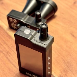 Op de afbeelding: Een zwarte en zilveren radardetector met een klein scherm en een zwarte en rode knop. De tekst "lazy punch" is zichtbaar op het apparaat. Het apparaat staat op een houten oppervlak.