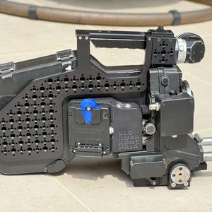 Puede incluir: Cámara de video profesional negra con asa, varios puertos y el texto "ELO KUVA KONE PAJA". La cámara tiene un panel lateral perforado y un soporte de batería azul. Diseñada para grabación de video profesional.