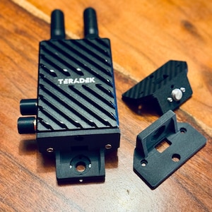 May include: A black Teradek wireless video transmitter with two antennas, a mounting bracket, and a small black accessory.