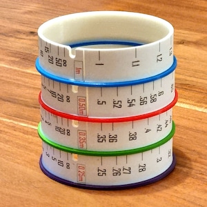 May include: A set of four white plastic measuring rings with different colored edges. The rings are marked with measurements in inches and centimeters. The rings are stacked on top of each other.