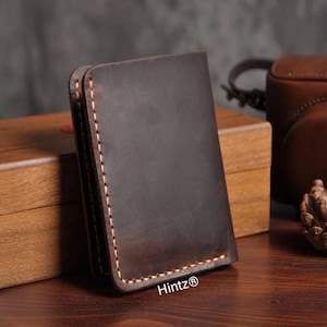 May include: A dark brown leather wallet with visible stitching along the edges. The wallet is rectangular and appears to be made of high-quality leather. The brand name "Hintz®" is visible on the bottom right corner.