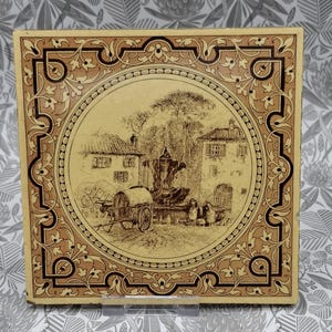 May include: A square ceramic tile featuring a central circular illustration of a village scene with a horse-drawn cart, a fountain, and buildings. The border is decorated with a brown and cream floral pattern with dark brown geometric lines.