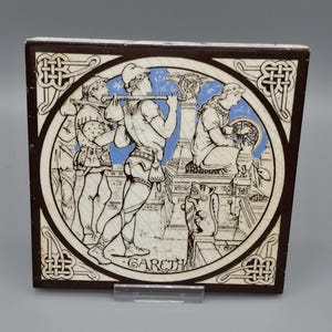 May include: Square ceramic tile with a brown border and a central circular design featuring a medieval scene in black and white with blue accents. The word "GARETH" is written at the bottom of the design.