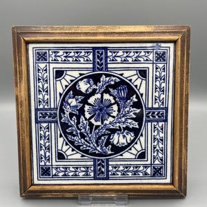 May include: Square ceramic tile with a floral design in a wooden frame. The tile features a central circular motif with a blue and white flower and leaf pattern. The frame is a light brown color.