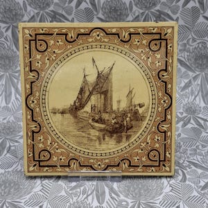 Op de afbeelding: Een vierkante keramische tegel met een sepia-kleurige illustratie van een zeilboot en een kleine roeiboot met figuren op het water. De centrale cirkelvormige afbeelding is omlijst door een decoratieve rand met bloemmotieven.