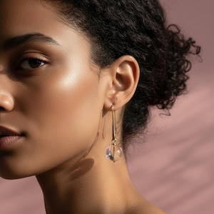May include: A close-up of a person wearing a gold-colored earring. The earring features a long, thin, gold bar with a clear, spherical bead at the bottom. The background is a soft pink.