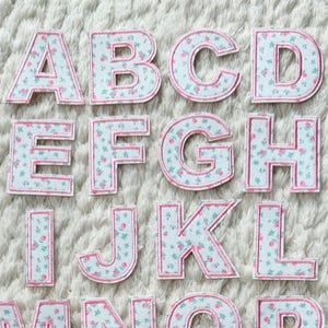 May include: A full set of fabric alphabet letters, each featuring a floral pattern in light blue and pink. The letters are outlined in pink and arranged on a white, textured surface. The letters are from A to Z.
