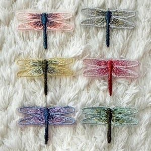 Könnte beinhalten: Sechs gestickte Libellen-Aufnäher, angeordnet in zwei Spalten. Die obere Reihe zeigt eine rosa und eine silberne Libelle. Die mittlere Reihe zeigt eine gelbe und eine rote Libelle. Die untere Reihe zeigt eine violette und eine hellgrüne Libelle.