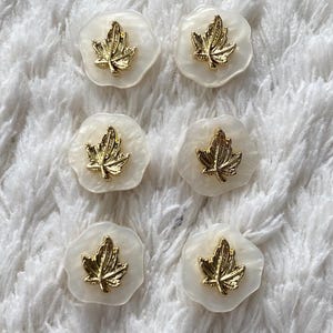 May include: Six white and gold leaf-shaped embellishments. Each embellishment features a gold-coloured maple leaf design set on a white, slightly translucent base. The items are arranged on a white, textured surface.