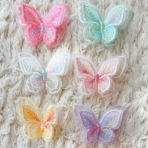 Op de afbeelding: Een verzameling van negen geborduurde vlinderpatches gerangschikt in rijen. De vlinders hebben verschillende pastelkleuren, waaronder roze, lichtblauw, wit, geel, paars, oranje en groen, met gedetailleerde draadstructuren op de vleugels.
