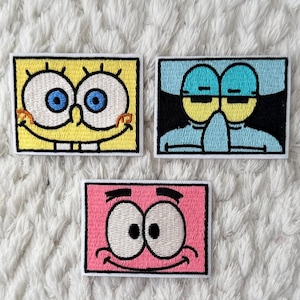 May include: Three embroidered patches featuring cartoon characters. The top patch is yellow with a smiling face and blue eyes. The middle patch is blue with a character's face. The bottom patch is pink with a smiling face.